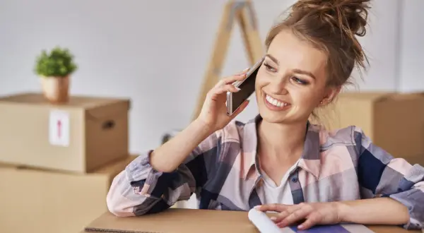 Uśmiechnięta kobieta rozmawia przez telefon, otoczona kartonami. Dowiaduje się, jak się zameldować w nowym miejscu.