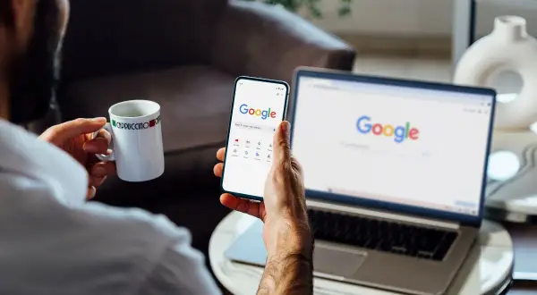 Mężczyzna z kubkiem kawy przegląda telefon, na którym widać stronę Google. Obok stoi laptop z otwartą wyszukiwarką. Chce wiedzieć, jak wylogować się z konta Google.
