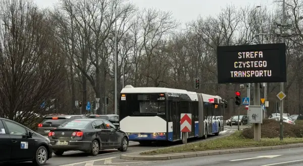 Autobus i samochody stoją w korku pod znakiem informującym o krakowskiej strefie czystego transportu.