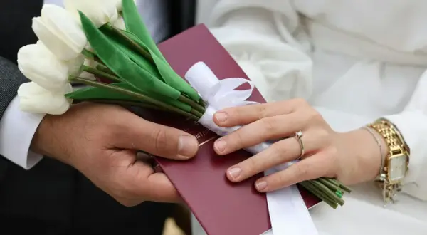 Dłoń mężczyzny i kobiety trzymają bukiet białych tulipanów i czerwony dokument.