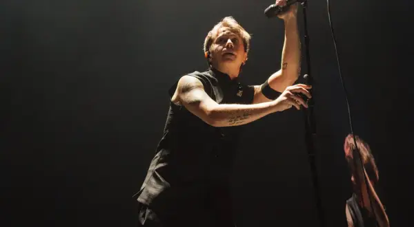 Lead singer of Nothing But Thieves, Conor Mason, belts into a microphone on a dark stage, his arm tattoos visible.