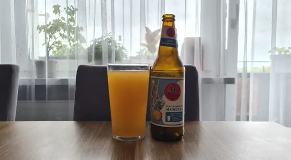 Szklanka z pomarańczowym napojem i butelka piwa bezalkoholowego mango ale na stole.