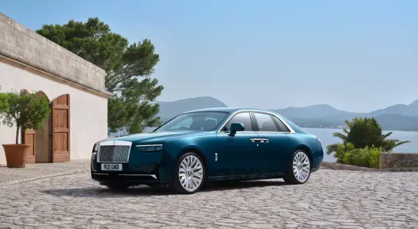 Elegancki Rolls-Royce Ghost w kolorze głębokiego turkusu zaparkowany na kamiennym dziedzińcu, z widokiem na morze i góry.