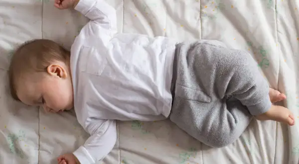 Un neonato non dorme, ma riposa sul fianco, vestito di bianco e grigio, su un lenzuolo con fiori stilizzati.