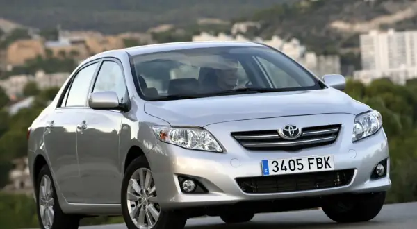Srebrna Toyota Corolla 2010 z widocznym kierowcą, zaparkowana na tle krajobrazu z budynkami.
