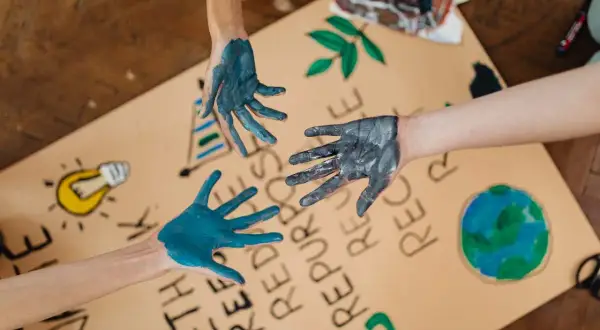 Manos pintadas de azul y gris sobre un cartel con dibujos de la Tierra, una bombilla y el símbolo de reciclaje. Un llamado a la acción para salvar el planeta.