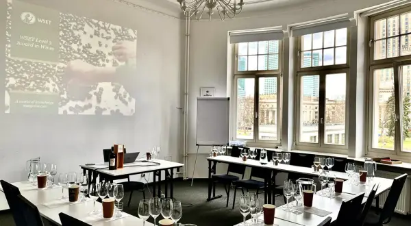 Sala do degustacji win, przygotowana do spotkania klubu wino. Na stołach kieliszki, dzbanki z wodą i butelka wina. Na ekranie logo WSET.