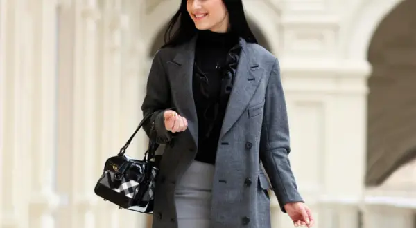 Uśmiechnięta it girl w eleganckim szarym płaszczu i z modną torebką, spacerująca po jasnym korytarzu.