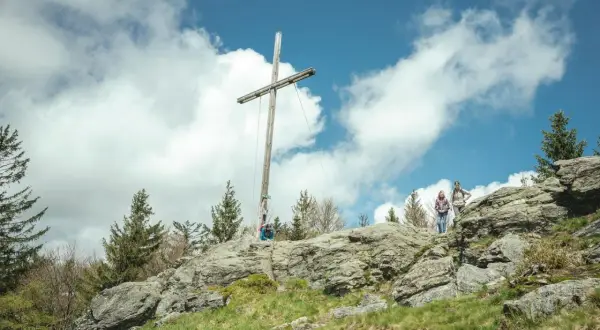 Wanderer auf einem felsigen Gipfel im Bayerischen Wald unter einem Holzkreuz. Ein typischer 8-Tausender-Ausblick.