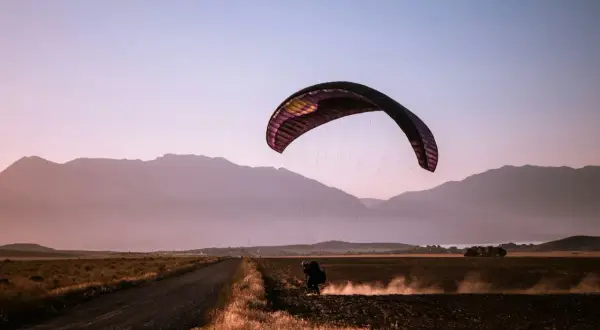 Parapentista aterrizando en un campo polvoriento al atardecer, rodeado de montañas. Una escena de turismo activo.