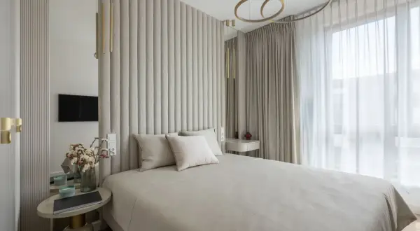 Elegancka sypialnia glamour w beżowych odcieniach. Miękkie, pikowane wezgłowie łóżka, złote akcenty i jasne zasłony tworzą przytulną atmosferę.