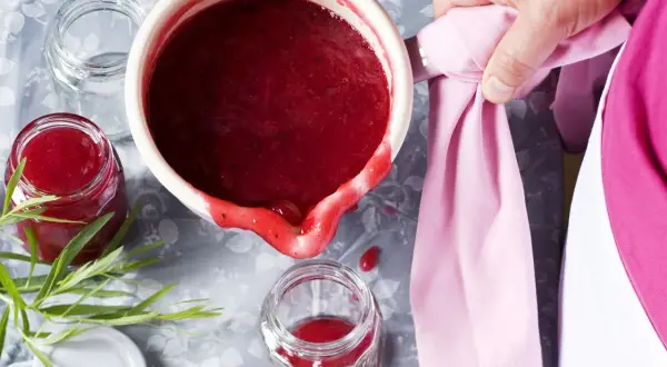 Frisch gekochte, einfache Johannisbeermarmelade wird in ein Glas gefüllt. Ein Hauch von Grün und ein rosa Tuch vervollständigen das Bild.