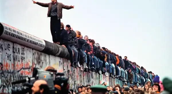 Tłum ludzi siedzi na Murze Berlińskim, świętując jego upadek. Jeden mężczyzna stoi na szczycie, wskazując palcem w dal.