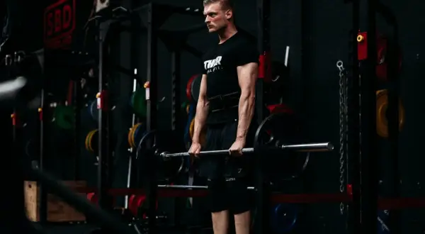 Ein Mann mit Bart und schwarzem T-Shirt hebt eine Langhantel im Fitnessstudio. Er trainiert seine Handkraft, um stärker zu werden.