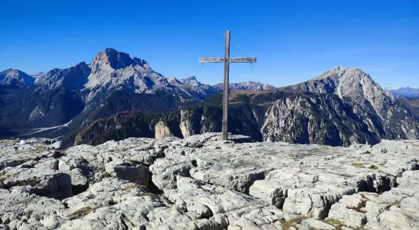 Holzkreuz auf felsigem Gipfel des Monte Piano, mit majestätischen Bergen im Hintergrund unter blauem Himmel.