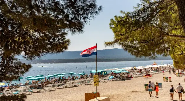 Chorwacka flaga powiewa nad pełną plażą z leżakami i parasolami. W tle błękitne morze i góry.