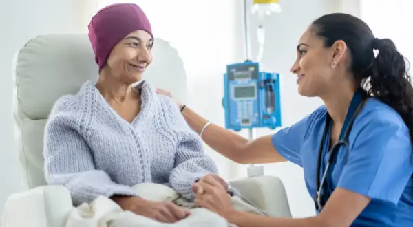 Eine Krankenschwester tröstet eine Patientin während des Chemotherapieablaufs. Die Patientin trägt eine lila Kopfbedeckung und eine Strickjacke.