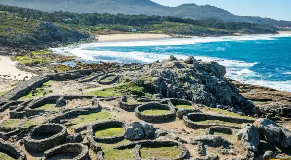 Ruinas circulares de piedra del castro de Baroña, con el mar azul y una playa de arena al fondo.