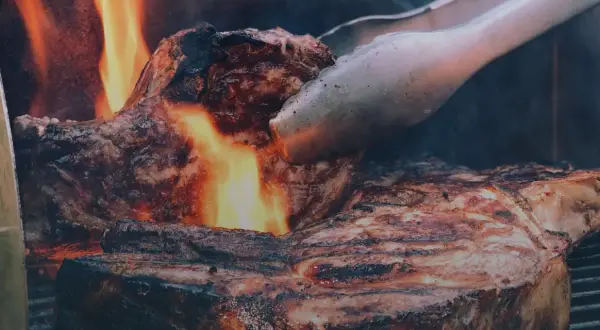 En saftig entrecote grillas över öppen eld, perfekt innertemp. Tången vänder köttet för en jämn stekyta.