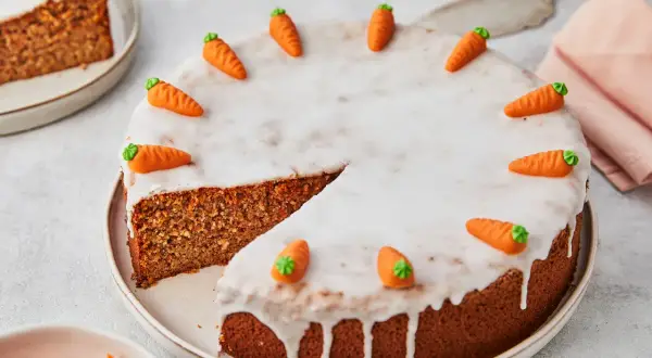 Ein saftiger karottenkuchen ohne mehl, verziert mit kleinen Marzipan-Karotten und Zuckerguss. Ein Stück ist bereits angeschnitten.