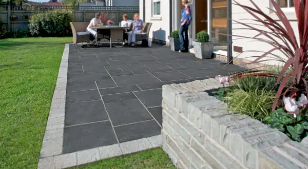 Un cortile moderno con eleganti piastrelle per cortile esterno grigie, bordate da un acciottolato chiaro. Persone rilassate al tavolo, circondati da verde.