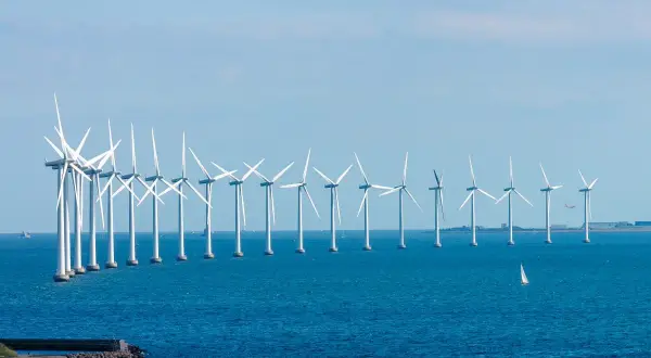 Rząd białych turbin wiatrowych tworzy imponującą farmę wiatrową na Bałtyku. Na horyzoncie widać żaglówkę i samolot.