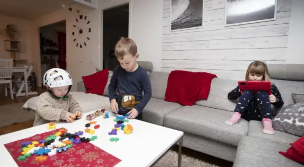 Tre barn leker med byggklossar och en surfplatta i ett vardagsrum. En pojke med hjälm bygger, en annan pojke leker med klossar, och en flicka tittar på en surfplatta. Leo Carmona barnen har kul.
