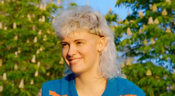 Ragazza con capelli biondi, corti sopra e lunghi dietro, stile anni '80, sorride all'aperto con alberi fioriti sullo sfondo.