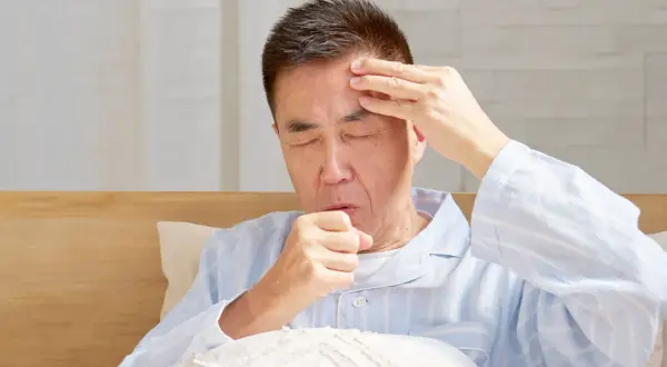Mann im Bett mit geschlossenen Augen, der sich die Stirn reibt und hustet. Er leidet unter Kopfschmerzen beim Husten.