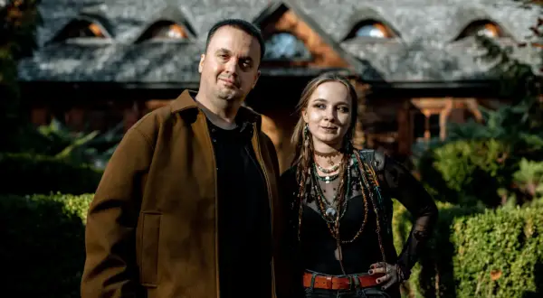 Para pozuje przed domem z kamiennym dachem. Mężczyzna w brązowej kurtce i kobieta w czarnej bluzce z wieloma naszyjnikami. Wyglądają jakby wyszli z planu filmu o straszydłach.