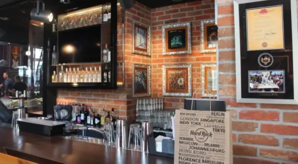 Wnętrze Hard Rock Cafe w Gdańsku: bar z butelkami alkoholu, ozdobne ramki na ścianie z cegły i torba z nazwami miast.