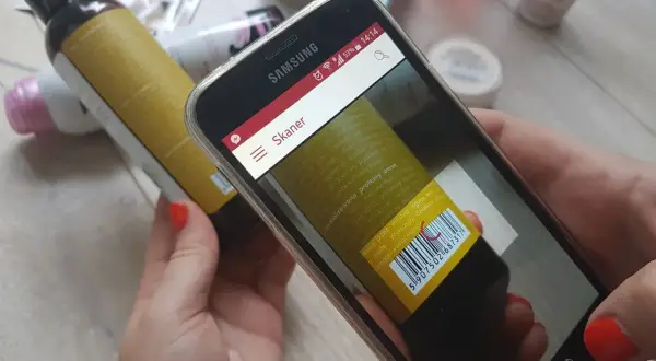 Ręce trzymają smartfon z otwartą aplikacją do sprawdzania składu kosmetyków. Na ekranie widoczny jest skaner kodu kreskowego skierowany na etykietę butelki.