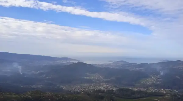 Vista panorámica desde el monte Galiñeiro, con un valle salpicado de casas y el mar al fondo bajo un cielo nublado.
