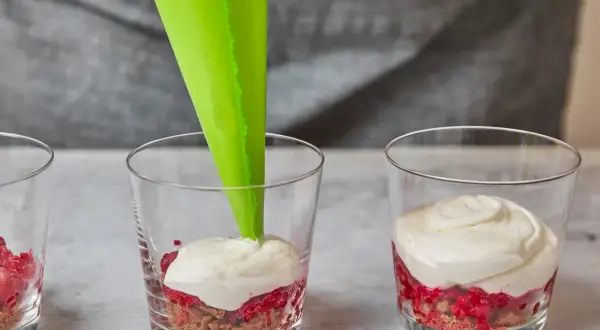 Cremige Masse wird mit Spritzbeutel in Gläser mit Himbeeren und Spekulatius-Bröseln gefüllt.