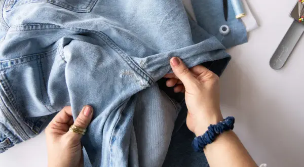 Hände reparieren ein Loch in Jeans mit einem Flicken. Nähutensilien liegen bereit.