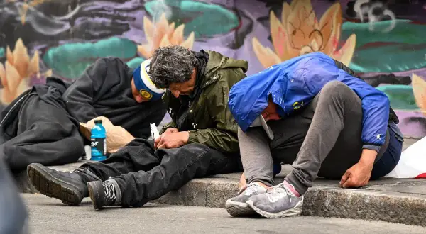 Trzech mężczyzn siedzi na chodniku, zgarbionych, z butelką napoju obok. Obraz ten może symbolizować problem uzależnień, w tym fentanyl w Polsce.