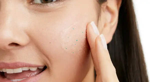 En kvinna applicerar en ansiktsskrubb med små, vita pormaskar-liknande korn för att exfoliera huden.