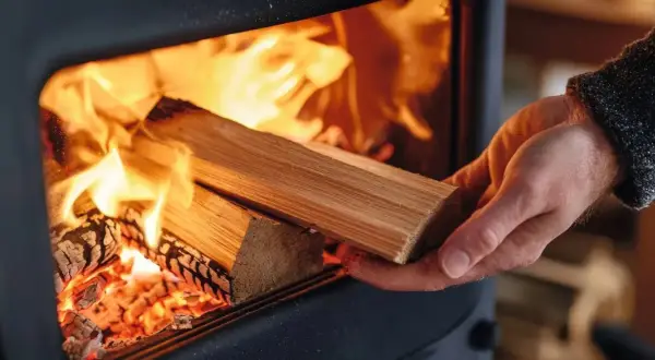 Eine Hand legt Holz in einen brennenden Kaminofen. Die Flammen lodern hell und wärmend.