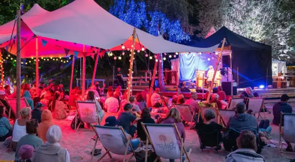 Menschen genießen ein **musik und stille festival** unter einem beleuchteten Zelt. Musiker spielen auf einer Bühne, während das Publikum auf Liegestühlen im Sand sitzt.
