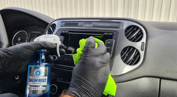 Ręce w czarnych rękawiczkach czyszczą deskę rozdzielczą samochodu, używając preparatu do regeneracji plastików.