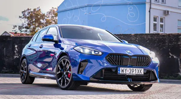 Błyszczące, niebieskie BMW 120 z czarnymi felgami stoi na brukowanym placu przed budynkiem z niebieską, ozdobną fasadą.