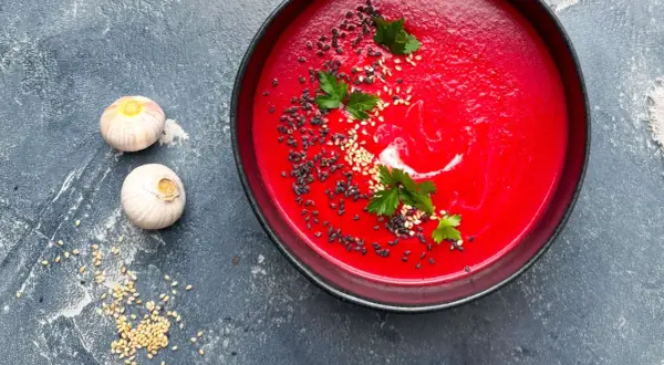 Eine leuchtend rote Bete Suppe, garniert mit Sesam und Petersilie, neben zwei Knoblauchzehen.
