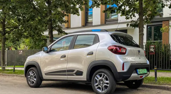 Srebrna Dacia Spring zaparkowana pod drzewami. Ten elektryczny samochód oferuje dobry zasięg na miejskie podróże.