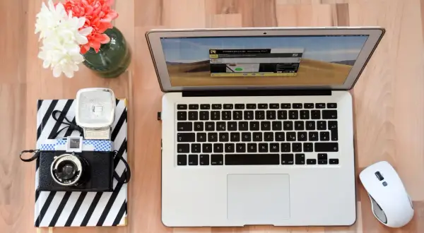 Jak zgrać zdjęcia z aparatu na laptop? Na biurku leży aparat, notatnik, laptop i myszka.