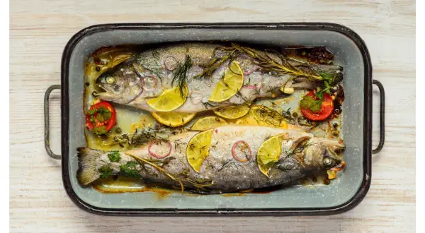 Deux truites au four, garnies de rondelles de citron, d'oignon rouge, de tomates cerises et d'herbes aromatiques, dans un plat en émail.