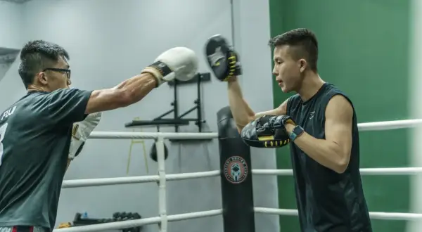 Two boxers train in a ring, one throwing a punch at the other's mitts during a reflex bag workout.