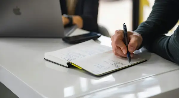 Dłoń pisząca długopisem w notatniku na białym biurku, obok laptopa.