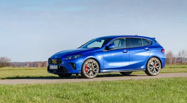 Błękitne BMW 120 na tle łąki i nieba. Sportowe felgi i czerwone zaciski hamulcowe dodają mu charakteru.