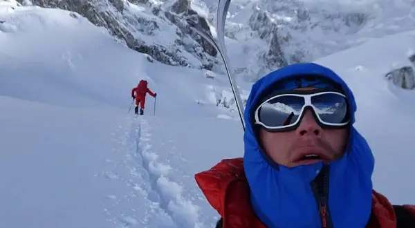 Tom Ballard im Schnee, mit Skiern auf dem Rücken, blickt in die Ferne. Ein weiterer Skifahrer ist im Hintergrund zu sehen.