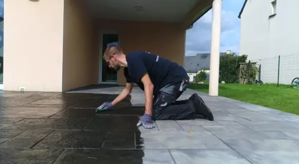 Un homme agenouillé effectue le nettoyage de pierre naturelle extérieur avec une éponge, redonnant son éclat à la terrasse.